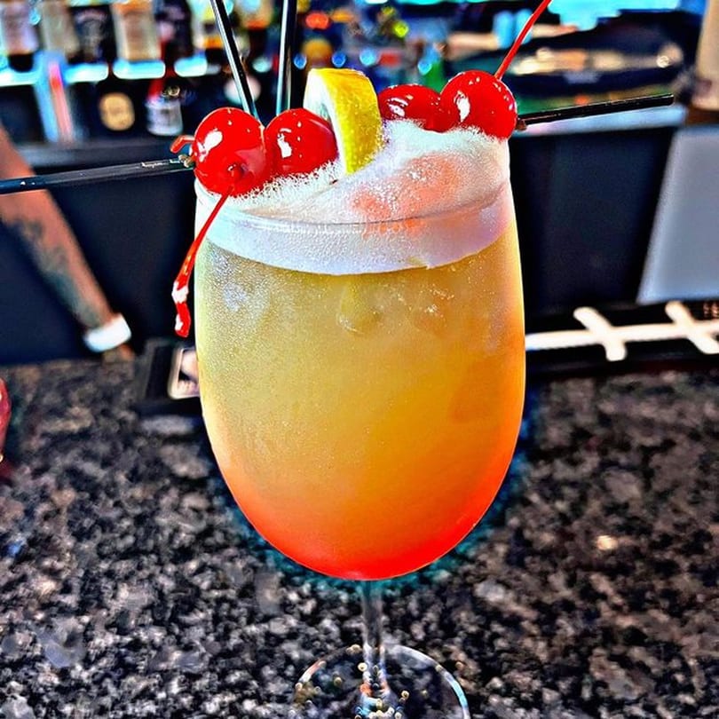 A vibrant yellow and orange cocktail with a foamy top, garnished with cherries and a lemon slice.