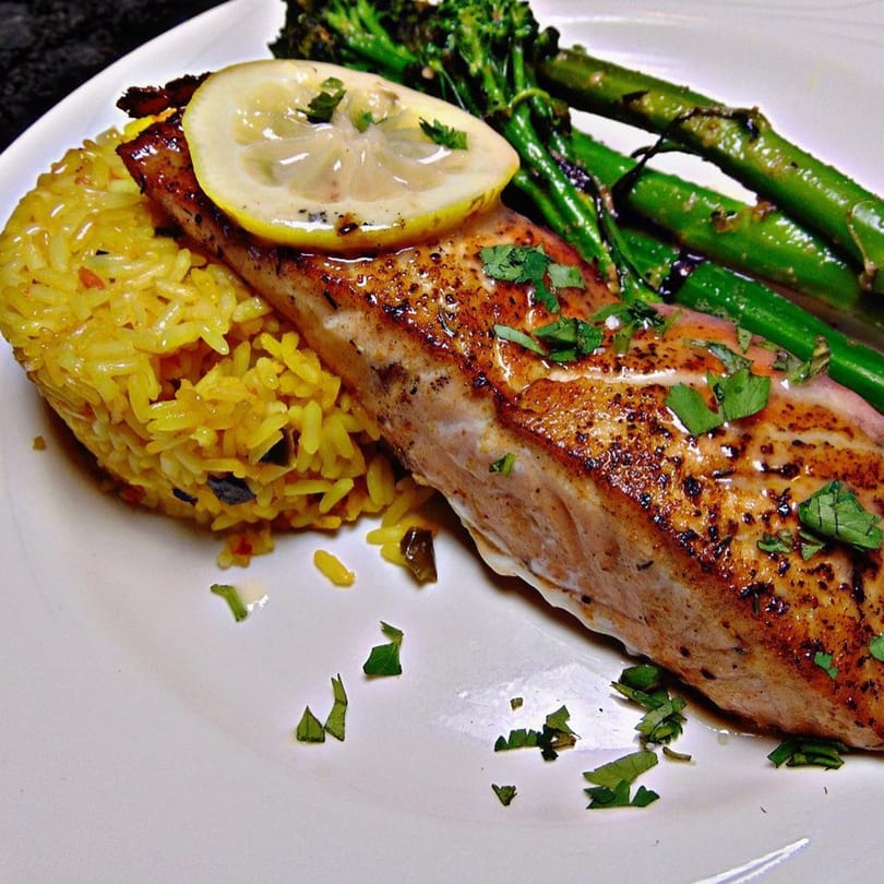 Grilled salmon with a lemon slice, yellow rice, and green asparagus on a white plate.